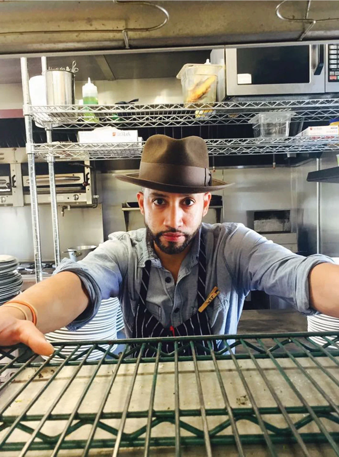 David de Castro in a brimmed hat and apron, leaning on a wire shelf in a restaurant kitchen