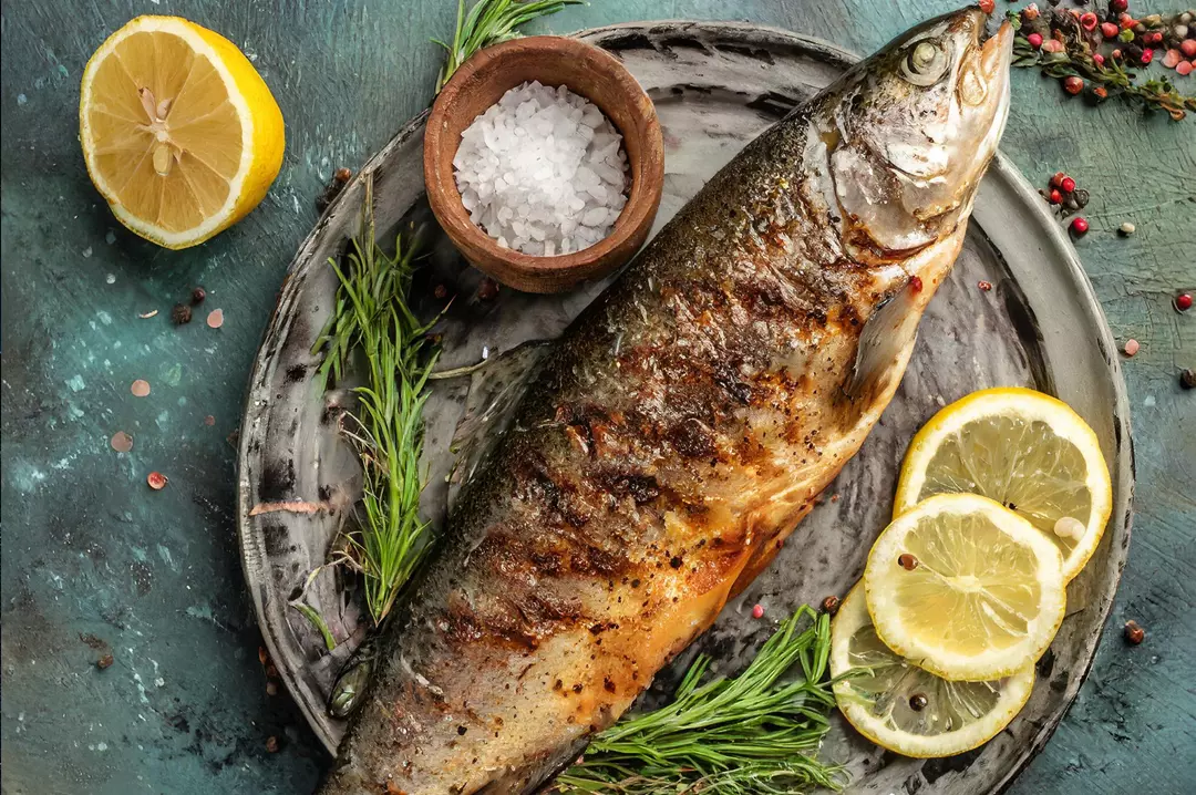 A whole roasted fish with lemon halves, herbs and a dish of salt on a dark tabletop