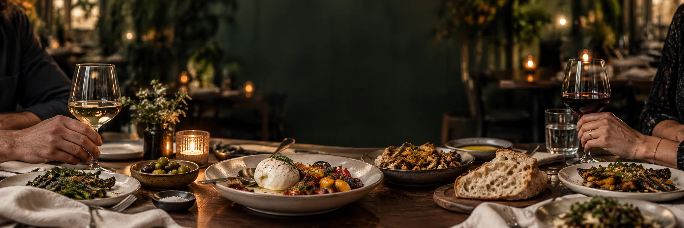 A shared bistro table with burrata, bread, olives and glasses of white and red wine by candlelight